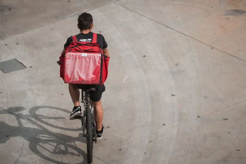 A food delivery cyclist rides on a concrete path with a red insulated backpack.